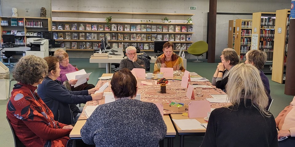 Rond een tafel met tafelkleed zitten mensen teksten te lezen en te luisteren